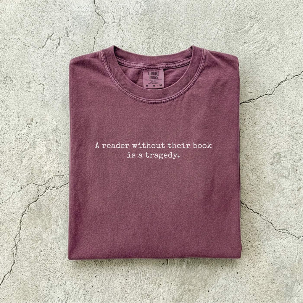 Berry Comfort Colors tee folded flat on a concrete background reading "A reader without their book is a tragedy." in a minimal typewriter font.