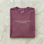 Berry Comfort Colors tee folded flat on a concrete background reading "A reader without their book is a tragedy." in a minimal typewriter font.