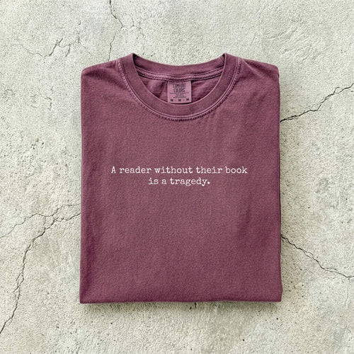 Berry Comfort Colors tee folded flat on a concrete background reading "A reader without their book is a tragedy." in a minimal typewriter font.