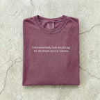 Berry Comfort Colors tee folded flat on a concrete background reading "Introverted, but willing to discuss spicy books." in a minimal typewriter font.