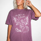 Woman wearing a berry color relaxed fit Comfort Colors tee with a vintage graphic of a woman warrior dragon rider and "We Ride at Dawn" text.