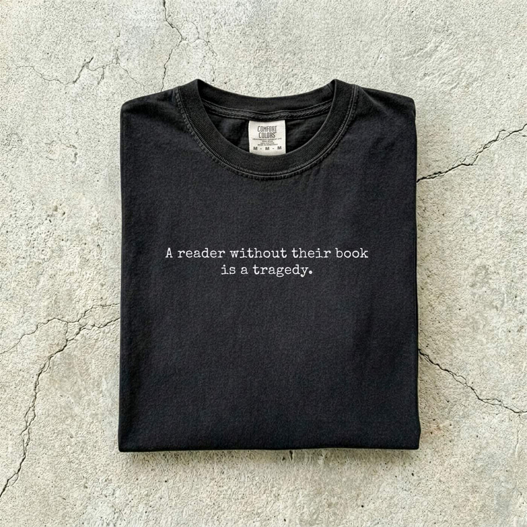 Black Comfort Colors tee folded flat on a concrete background reading "A reader without their book is a tragedy." in a minimal typewriter font.