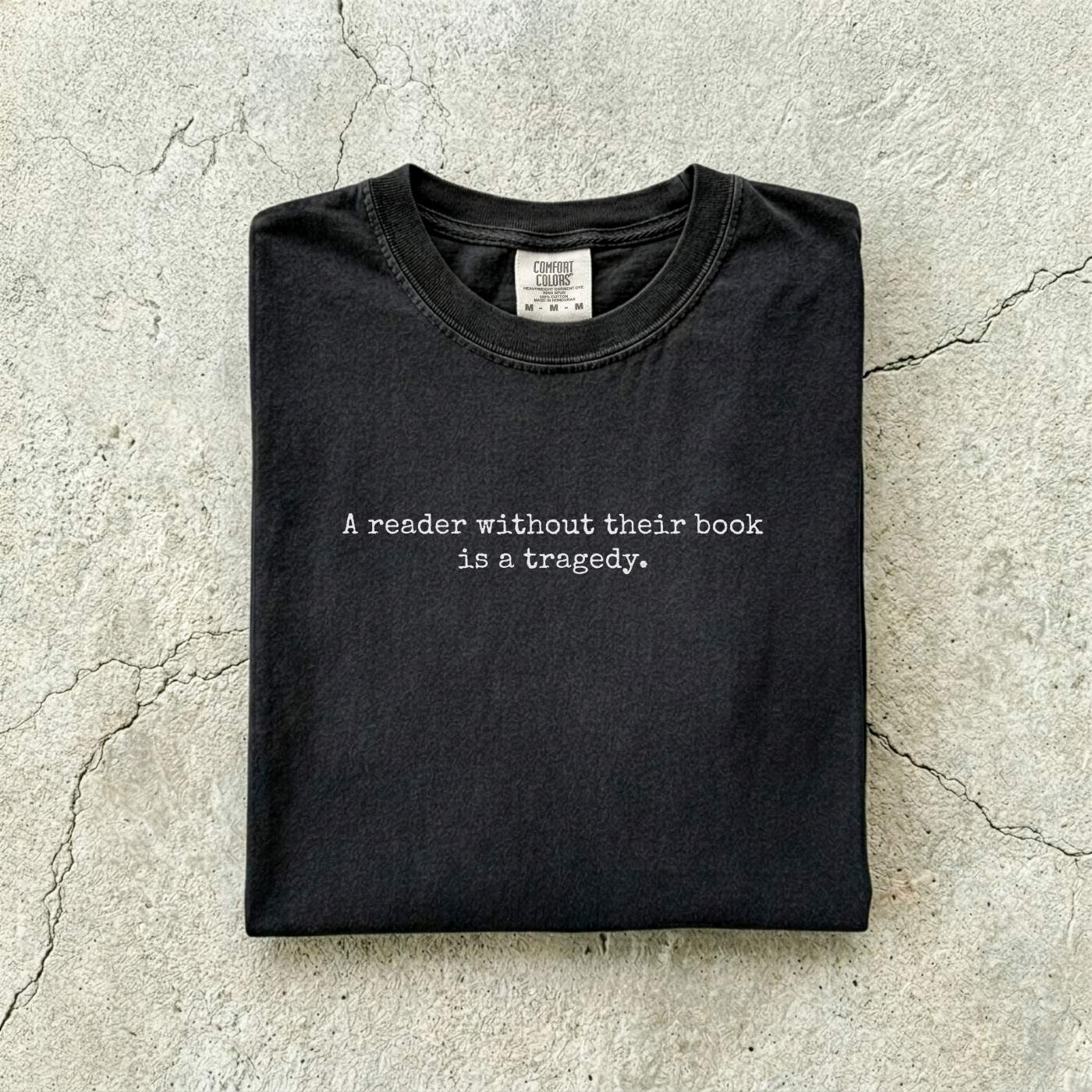 Black Comfort Colors tee folded flat on a concrete background reading "A reader without their book is a tragedy." in a minimal typewriter font.