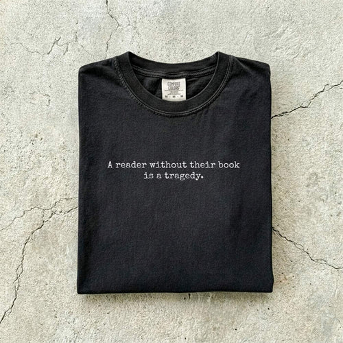 Black Comfort Colors tee folded flat on a concrete background reading "A reader without their book is a tragedy." in a minimal typewriter font.