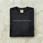 Black Comfort Colors tee folded flat on a concrete background reading "Enemies make the best lovers." in a minimal typewriter font.