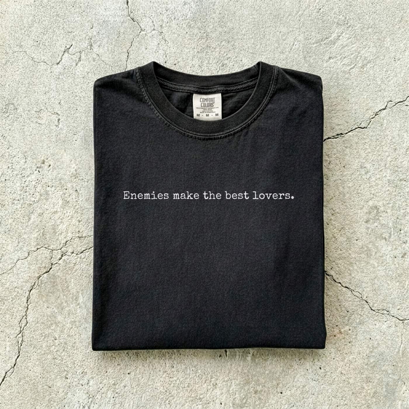 Black Comfort Colors tee folded flat on a concrete background reading "Enemies make the best lovers." in a minimal typewriter font.