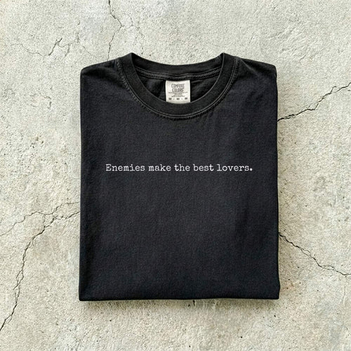 Black Comfort Colors tee folded flat on a concrete background reading "Enemies make the best lovers." in a minimal typewriter font.