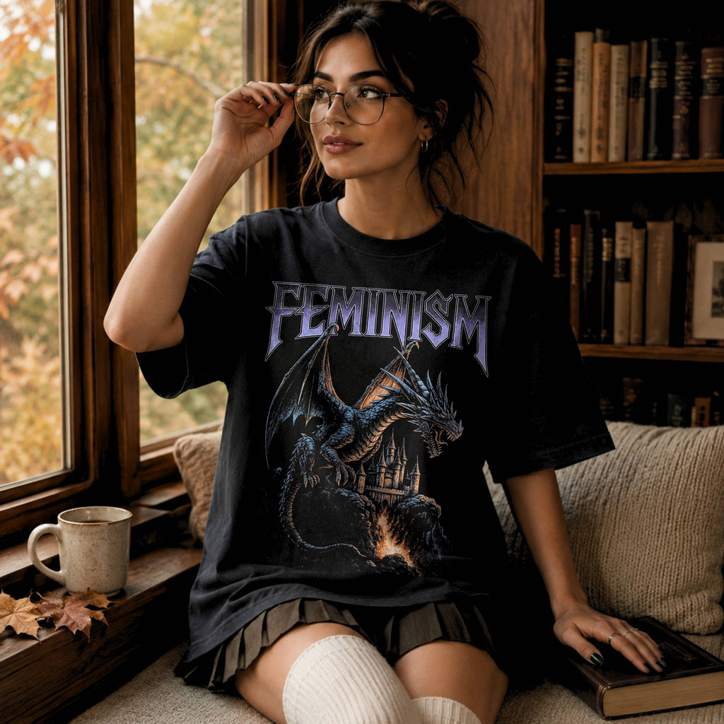 Woman wearing a black Comfort Colors relaxed tee with Feminism vintage metal graphic — dragon and lava-flowing castle design. She sits on a cozy couch with a bookcase behind her and looks out the library window.