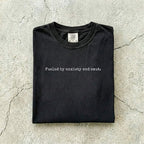 Black Comfort Colors tee folded flat on a concrete background reading "Fueled by anxiety and smut." in minimal typewriter font.