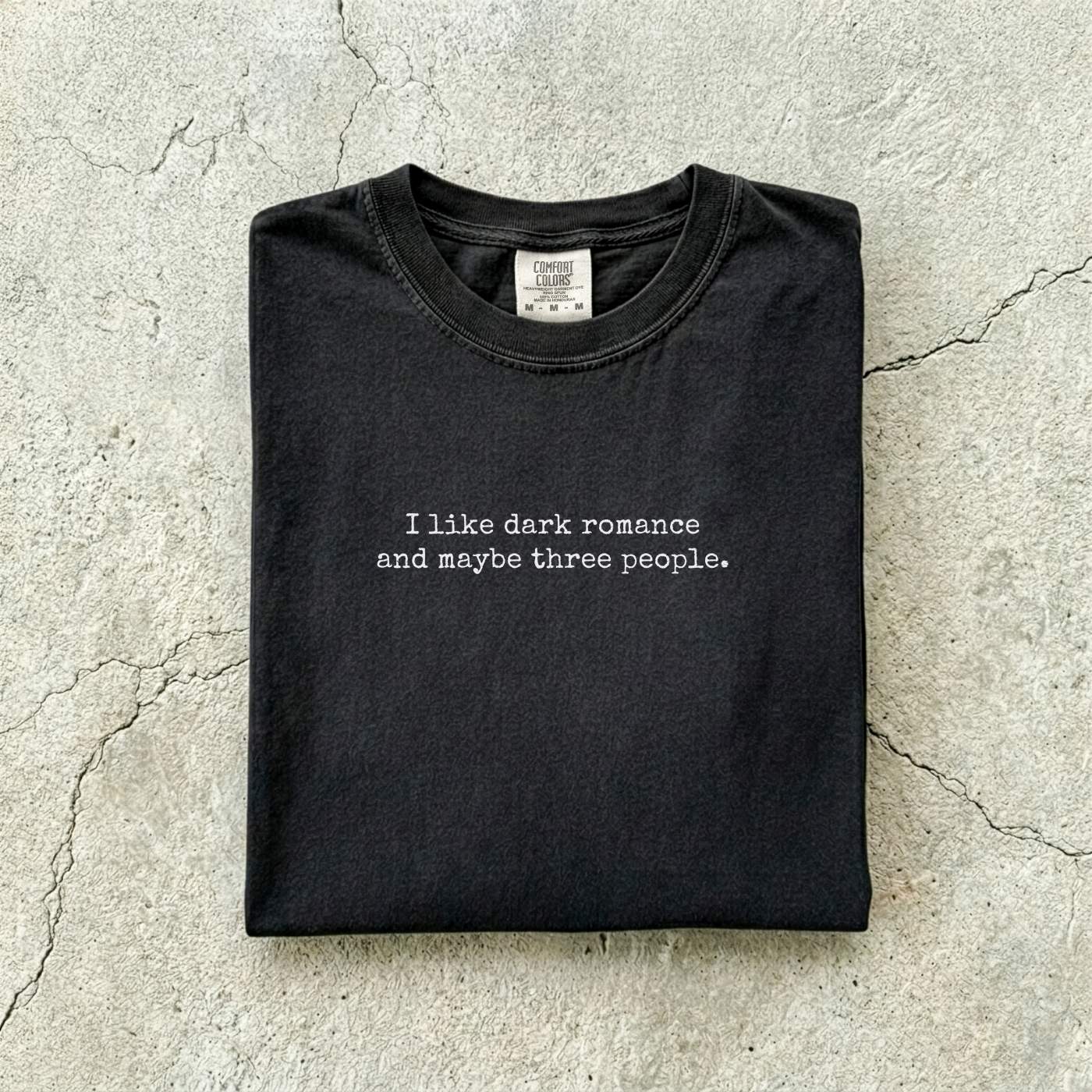 Black Comfort Colors tee folded flat reading "I like dark romance and maybe three people." in a minimalist typewriter font.