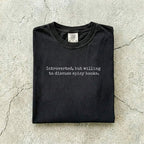 Black Comfort Colors tee folded flat on a concrete background reading "Introverted, but willing to discuss spicy books." in a minimal typewriter font.