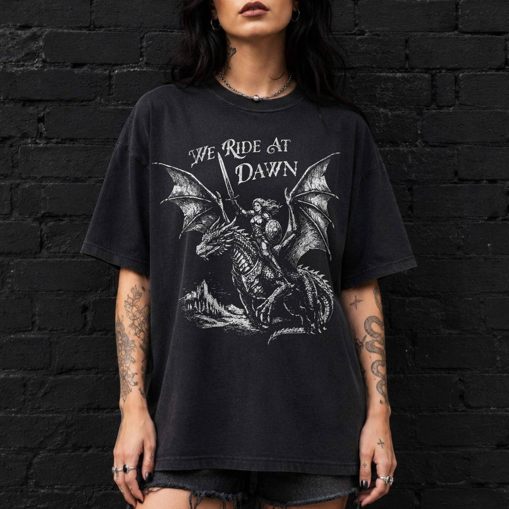 Woman wearing a black color relaxed fit Comfort Colors tee with a vintage graphic of a woman warrior dragon rider and "We Ride at Dawn" text.