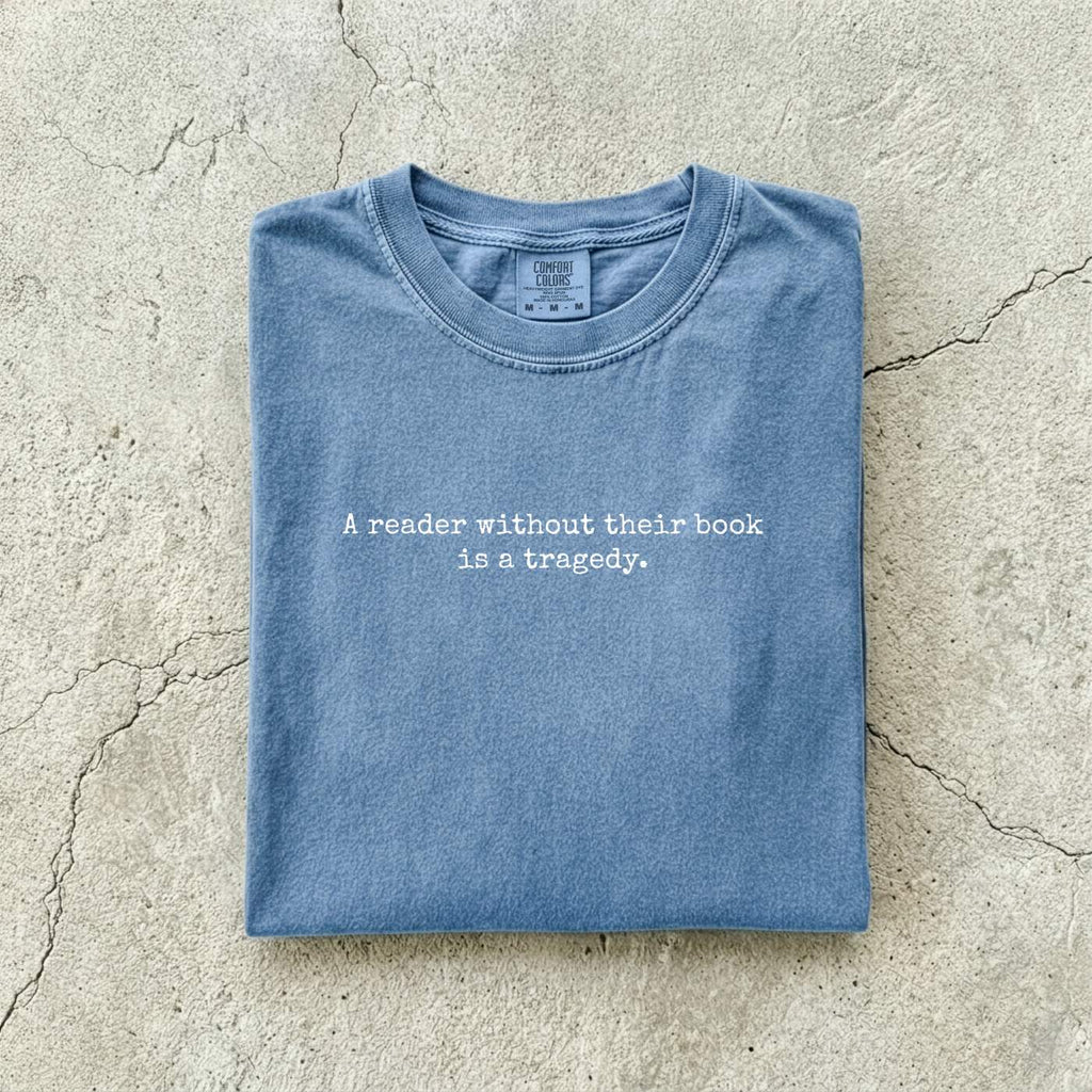 Blue Jean Comfort Colors tee folded flat on a concrete background reading "A reader without their book is a tragedy." in a minimal typewriter font.