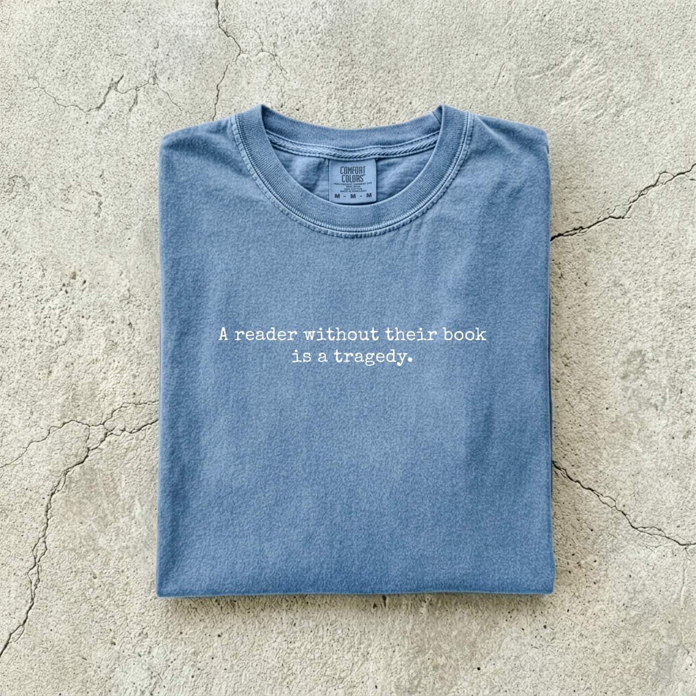 Blue Jean Comfort Colors tee folded flat on a concrete background reading "A reader without their book is a tragedy." in a minimal typewriter font.