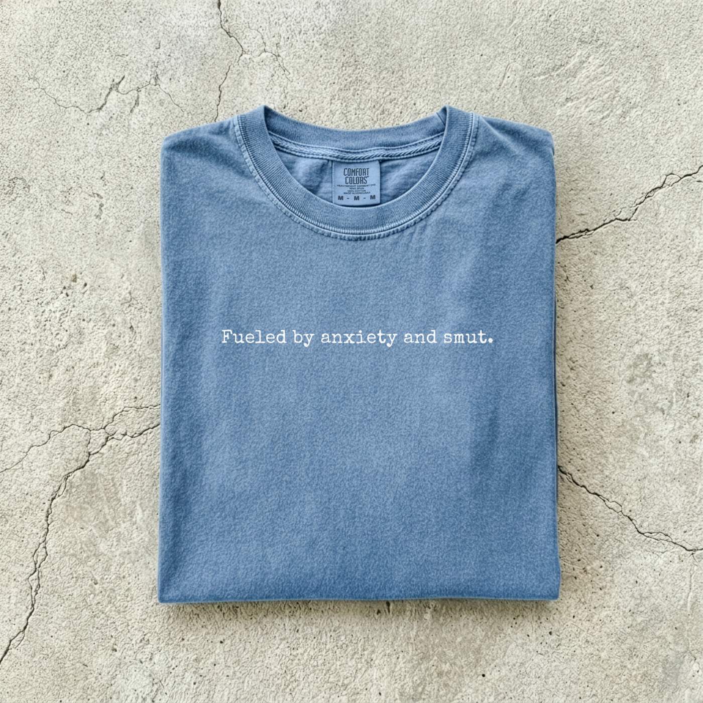 Blue Jean Comfort Colors tee folded flat on a concrete background reading "Fueled by anxiety and smut." in minimal typewriter font.