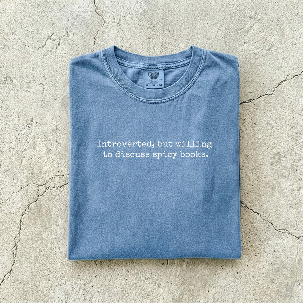 Blue Jean Comfort Colors tee folded flat on a concrete background reading "Introverted, but willing to discuss spicy books." in a minimal typewriter font.