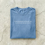 Blue Jean Comfort Colors tee folded flat on a concrete background reading "Introverted, but willing to discuss spicy books." in a minimal typewriter font.