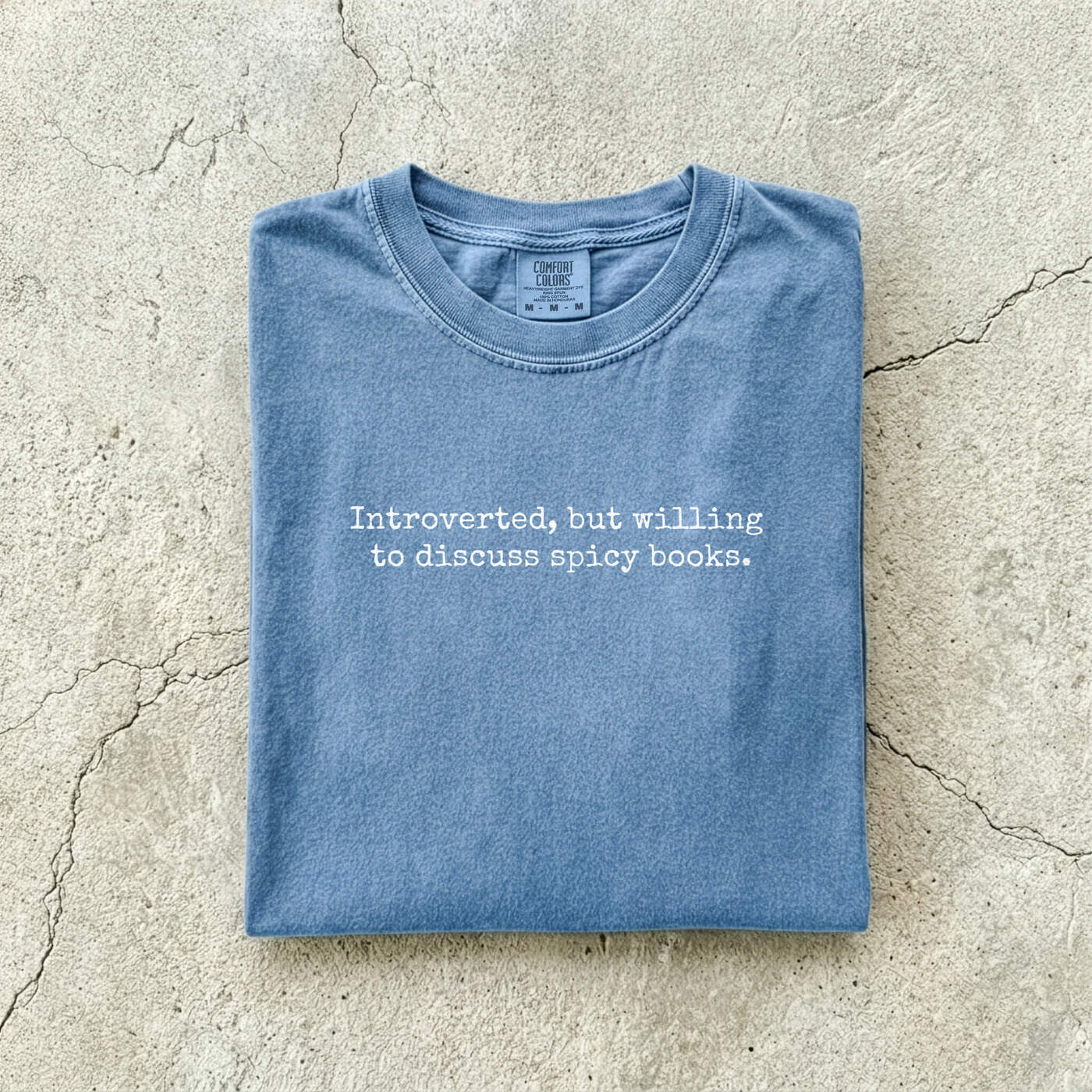 Blue Jean Comfort Colors tee folded flat on a concrete background reading "Introverted, but willing to discuss spicy books." in a minimal typewriter font.