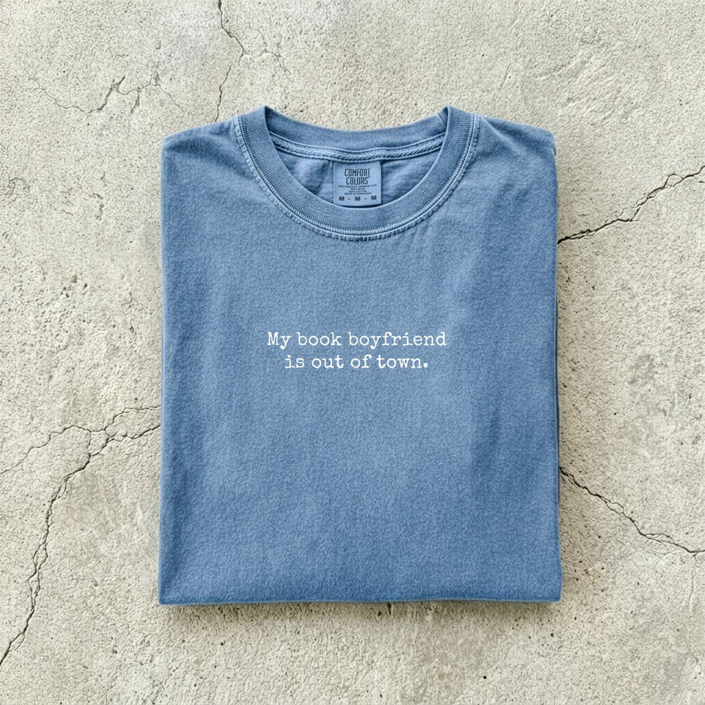 Blue Jean Comfort Colors tee folded flat on a concrete background reading "My book boyfriend is out of town." in a minimal typewriter font.