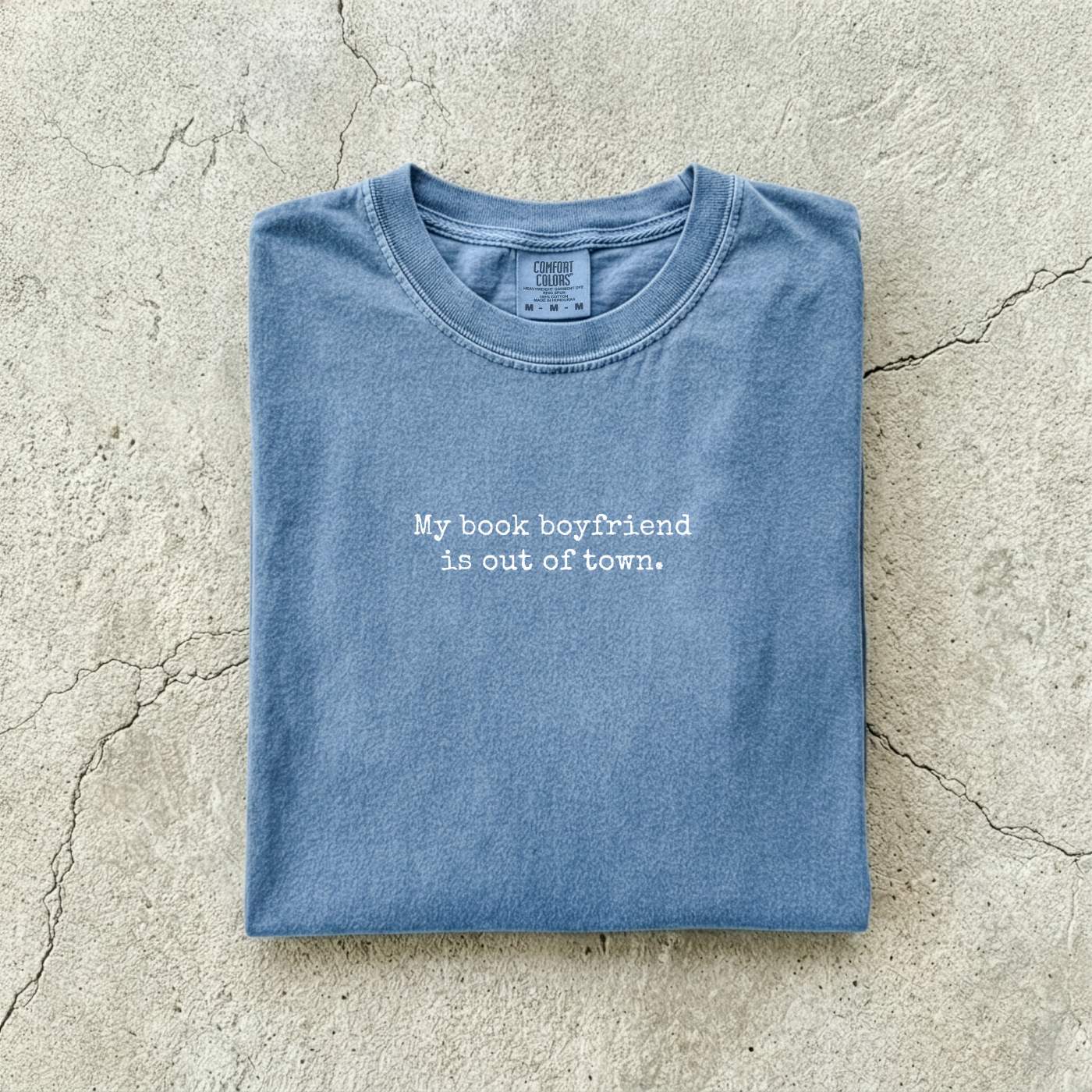 Blue Jean Comfort Colors tee folded flat on a concrete background reading "My book boyfriend is out of town." in a minimal typewriter font.