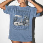 Blue Jean Comfort Colors 1717 relaxed fit t-shirt with gothic dark romance graphic — a raven perched on a skull, a snake coiled on stacked books, a candle, and florals, with the text "Dark Romance Book Club — Enter for the Plot, Stay for the Trauma"