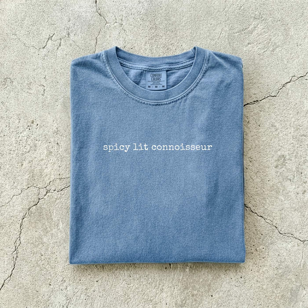 Blue Jean Comfort Colors tee folded flat on a concrete background reading "spicy lit connoisseur" in a minimal typewriter font.