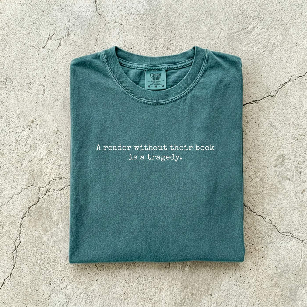 Blue Spruce Comfort Colors tee folded flat on a concrete background reading "A reader without their book is a tragedy." in a minimal typewriter font.