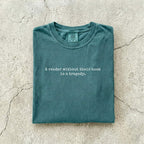 Blue Spruce Comfort Colors tee folded flat on a concrete background reading "A reader without their book is a tragedy." in a minimal typewriter font.