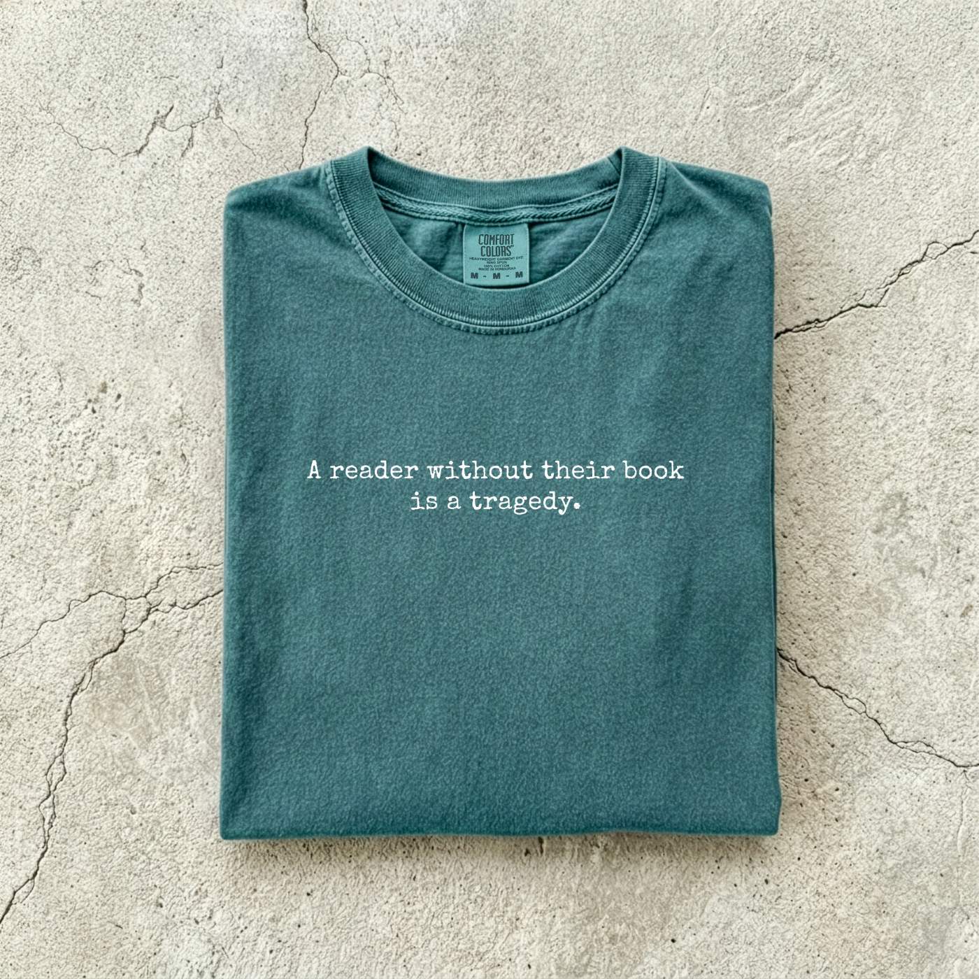 Blue Spruce Comfort Colors tee folded flat on a concrete background reading "A reader without their book is a tragedy." in a minimal typewriter font.