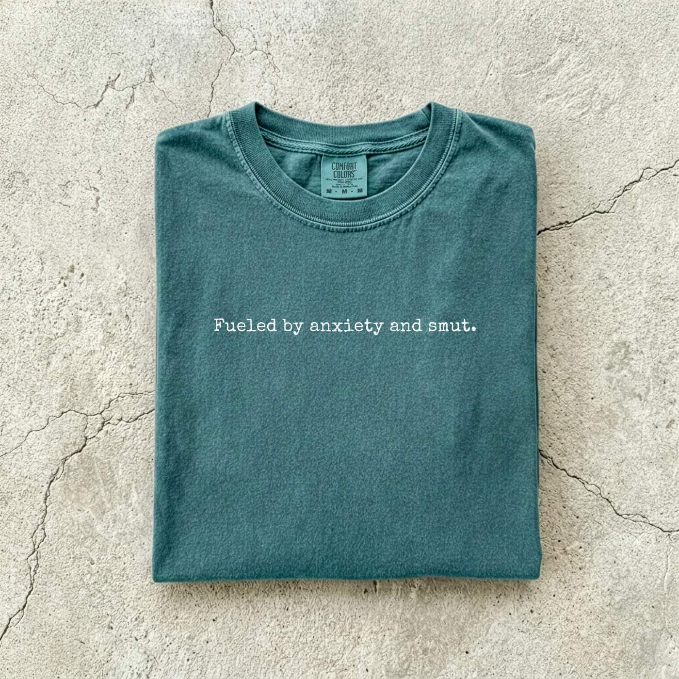 Blue Spruce Comfort Colors tee folded flat on a concrete background reading "Fueled by anxiety and smut." in minimal typewriter font.