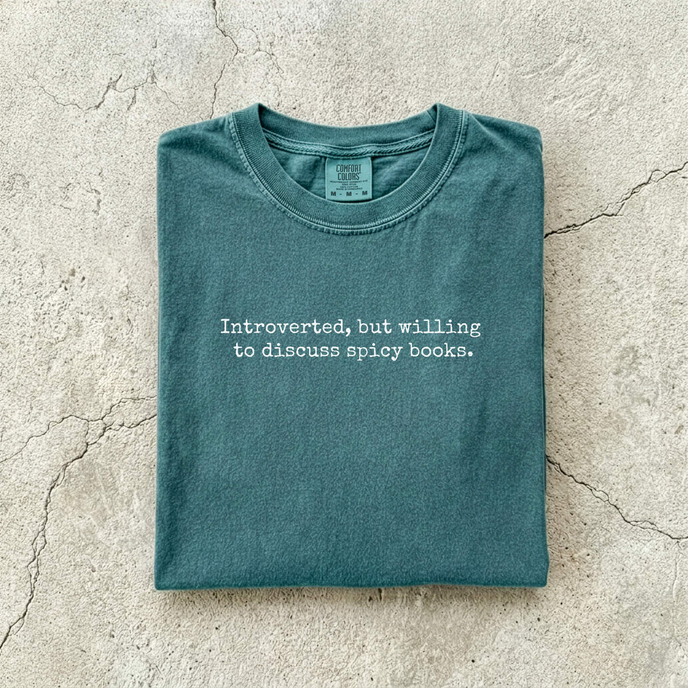 Blue Spruce Comfort Colors tee folded flat on a concrete background reading "Introverted, but willing to discuss spicy books." in a minimal typewriter font.
