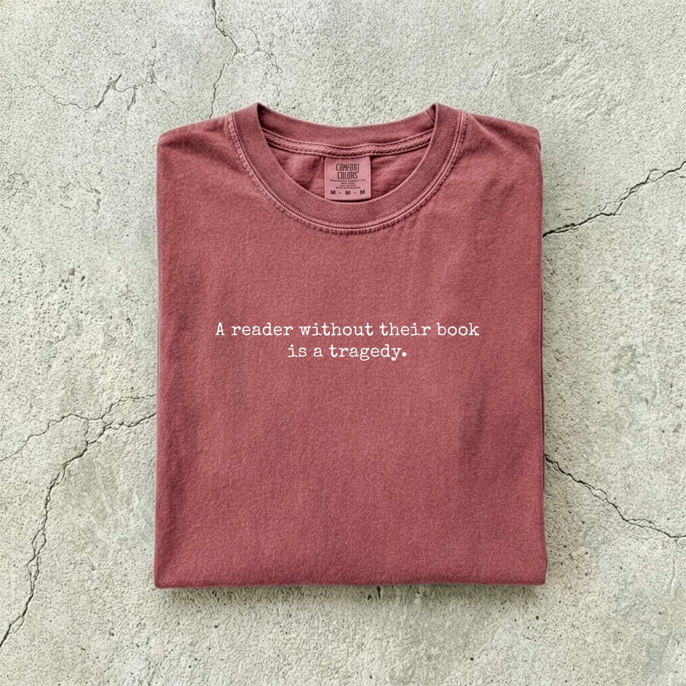 Brick Comfort Colors tee folded flat on a concrete background reading "A reader without their book is a tragedy." in a minimal typewriter font.