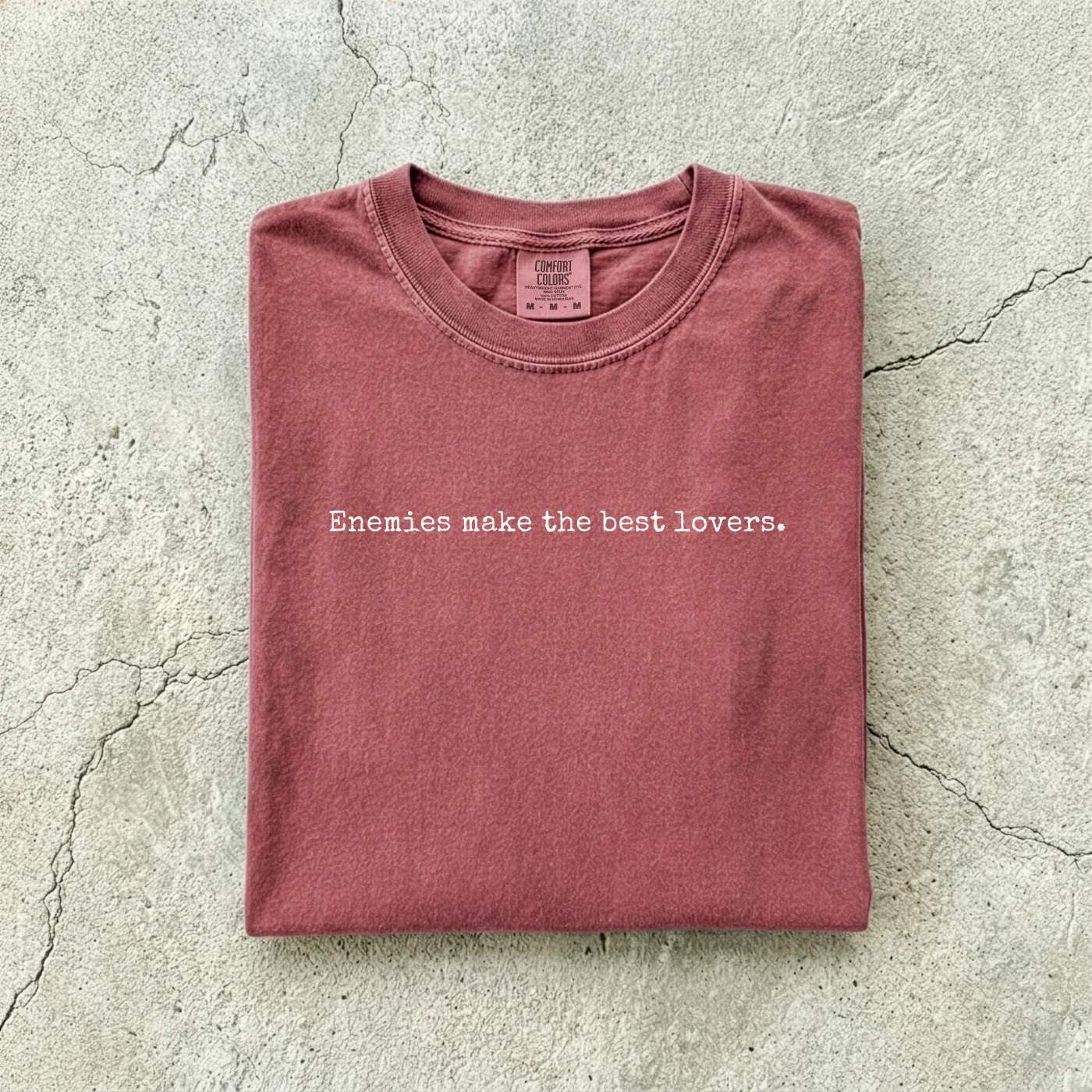 Brick Comfort Colors tee folded flat on a concrete background reading "Enemies make the best lovers." in a minimal typewriter font.