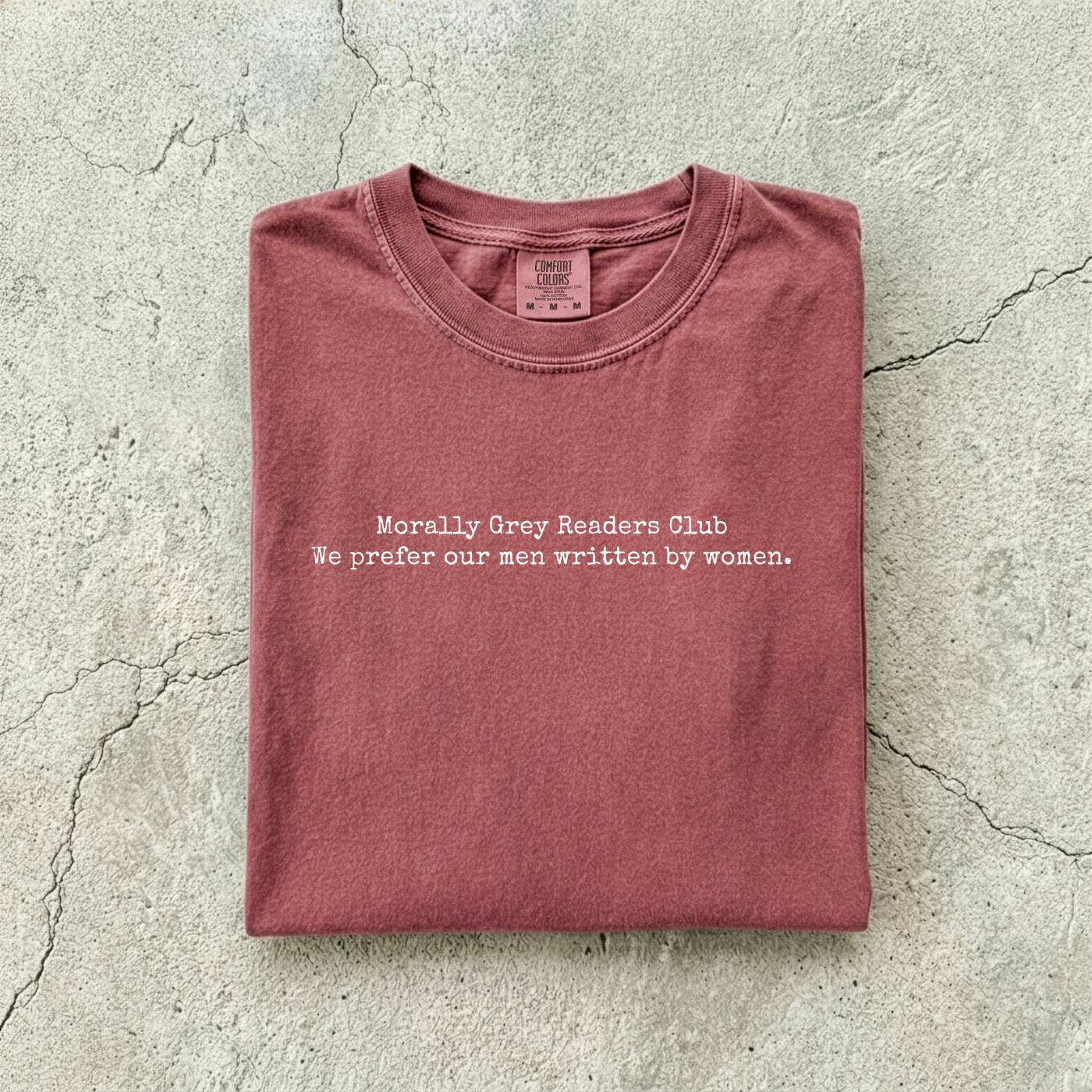 Brick Comfort Colors tee folded flat on a concrete background reading "Morally grey readers club. We prefer our men written by women." in a minimal typewriter font.