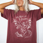 Woman wearing a brick color relaxed fit Comfort Colors tee with a vintage graphic of a woman warrior dragon rider and "We Ride at Dawn" text.