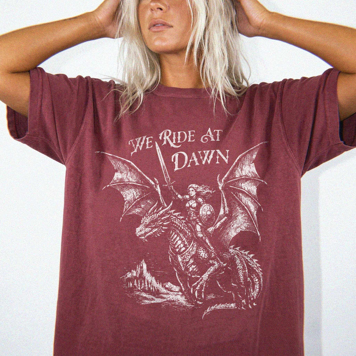 Woman wearing a brick color relaxed fit Comfort Colors tee with a vintage graphic of a woman warrior dragon rider and "We Ride at Dawn" text.
