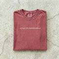 Brick Comfort Colors tee folded flat on a concrete background reading "spicy lit connoisseur" in a minimal typewriter font.