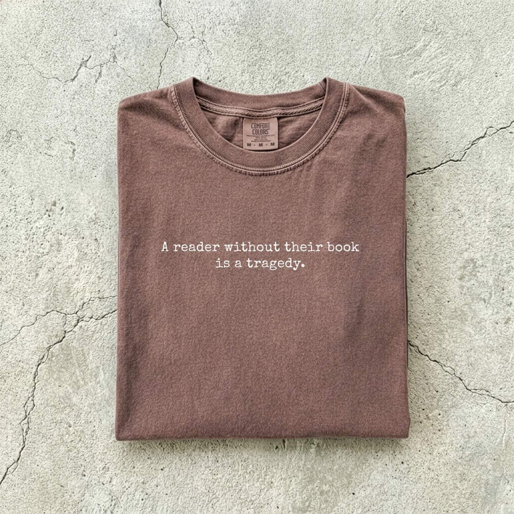 Espresso Comfort Colors tee folded flat on a concrete background reading "A reader without their book is a tragedy." in a minimal typewriter font.