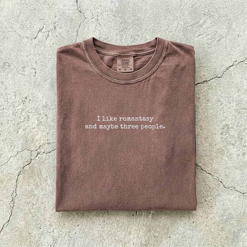 Espresso Comfort Colors tee folded flat reading "I like romantasy and maybe three people." in a typewriter font.