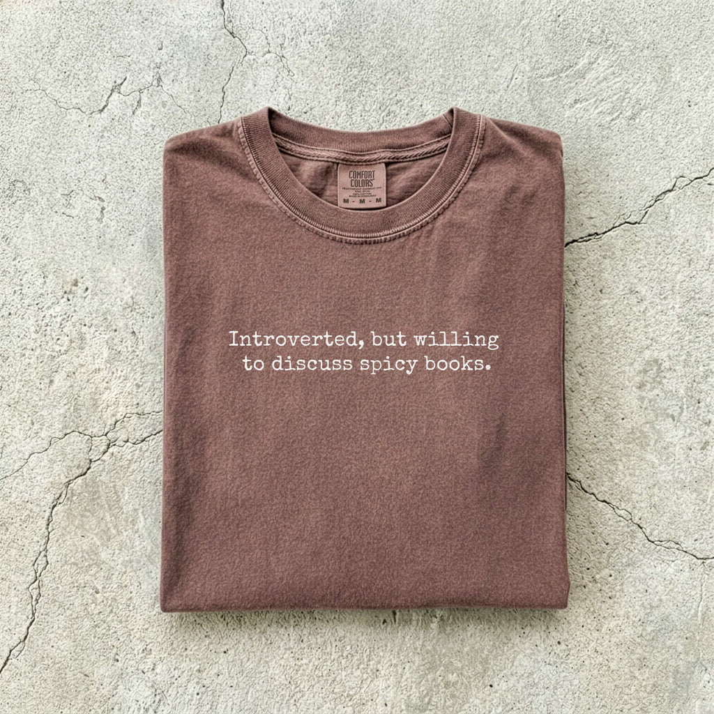 Espresso Comfort Colors tee folded flat on a concrete background reading "Introverted, but willing to discuss spicy books." in a minimal typewriter font.