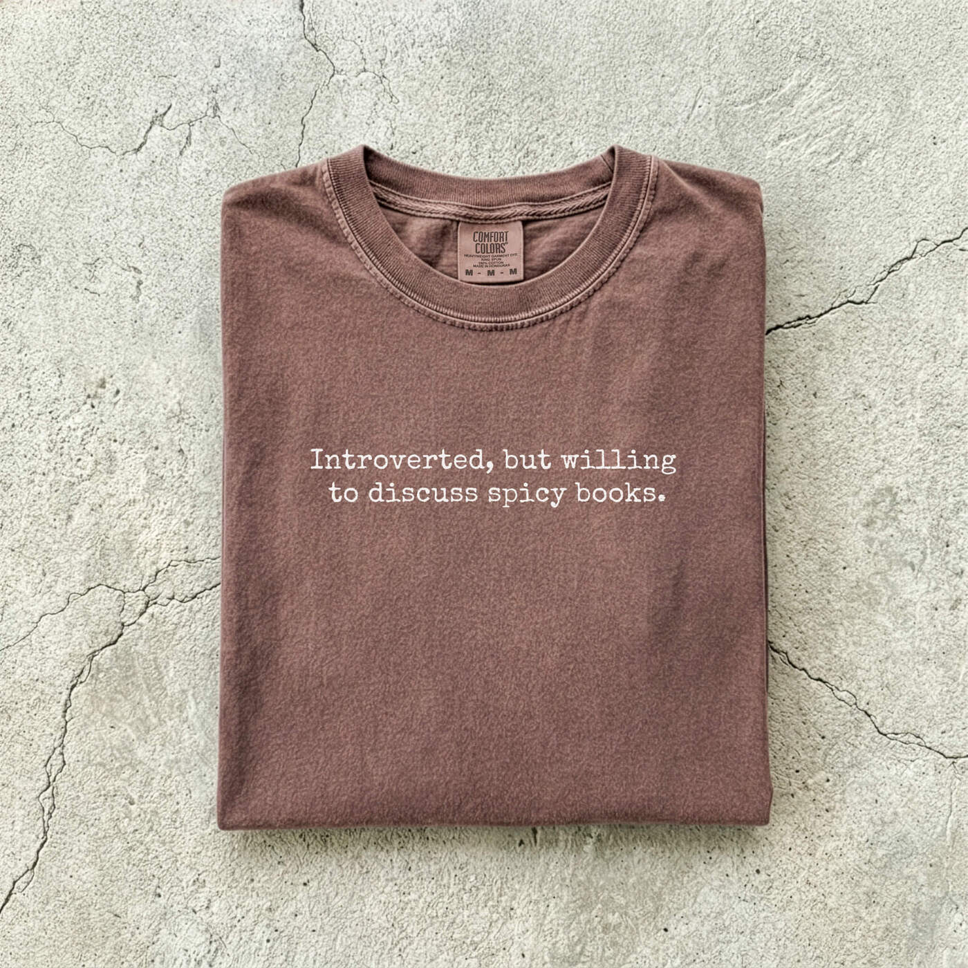 Espresso Comfort Colors tee folded flat on a concrete background reading "Introverted, but willing to discuss spicy books." in a minimal typewriter font.