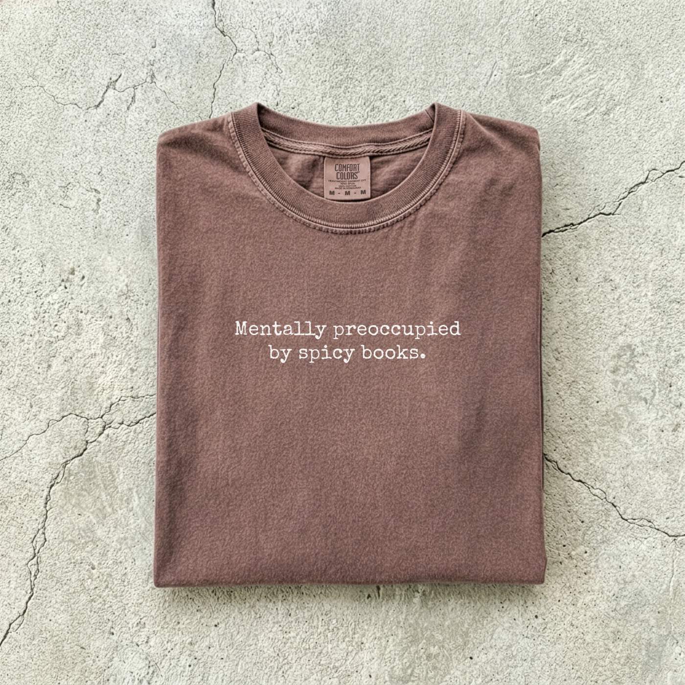 Espresso Comfort Colors tee folded flat on a concrete background reading "Mentally preoccupied by spicy books." in a minimalist typewriter font.