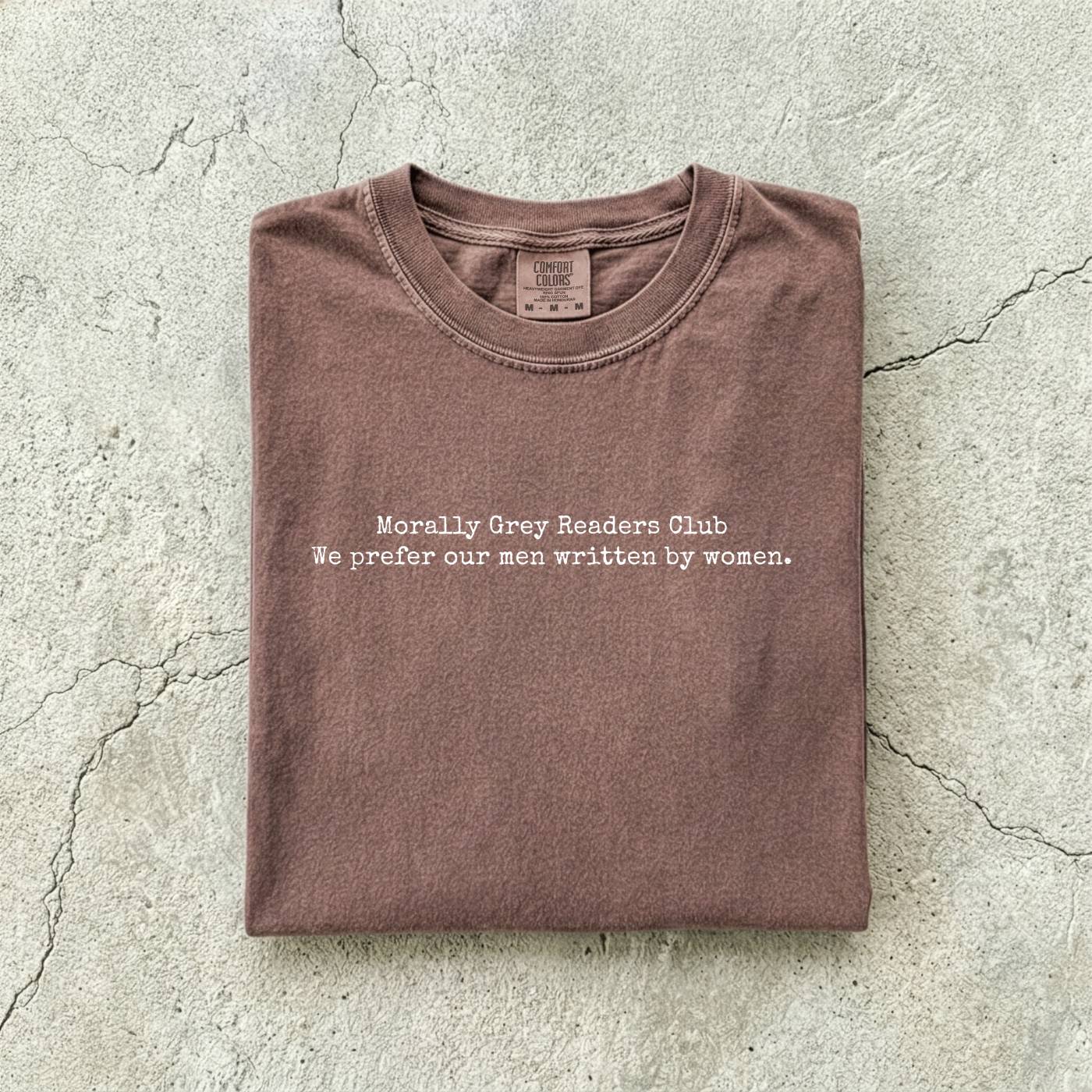 Espresso Comfort Colors tee folded flat on a concrete background reading "Morally grey readers club. We prefer our men written by women." in a minimal typewriter font.