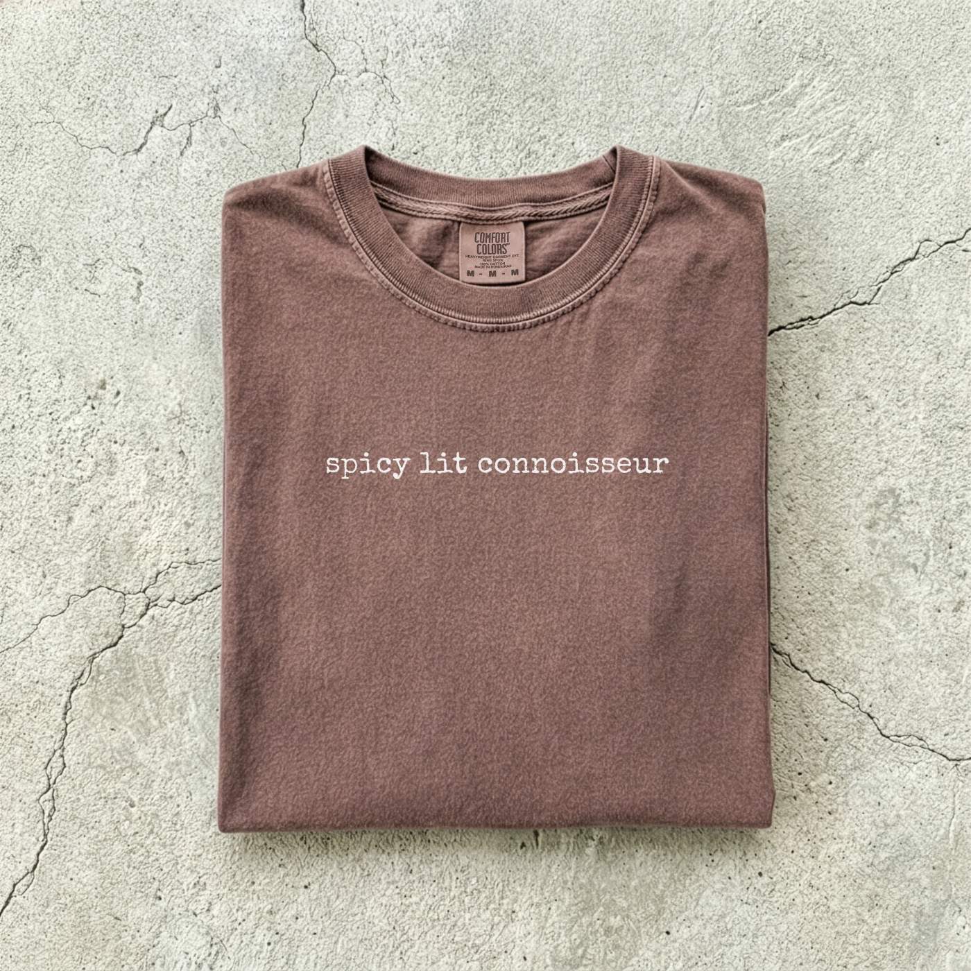 Espresso Comfort Colors tee folded flat on a concrete background reading "spicy lit connoisseur" in a minimal typewriter font.