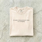 Ivory Comfort Colors tee folded flat on a concrete background reading "A reader without their book is a tragedy." in a minimal typewriter font.