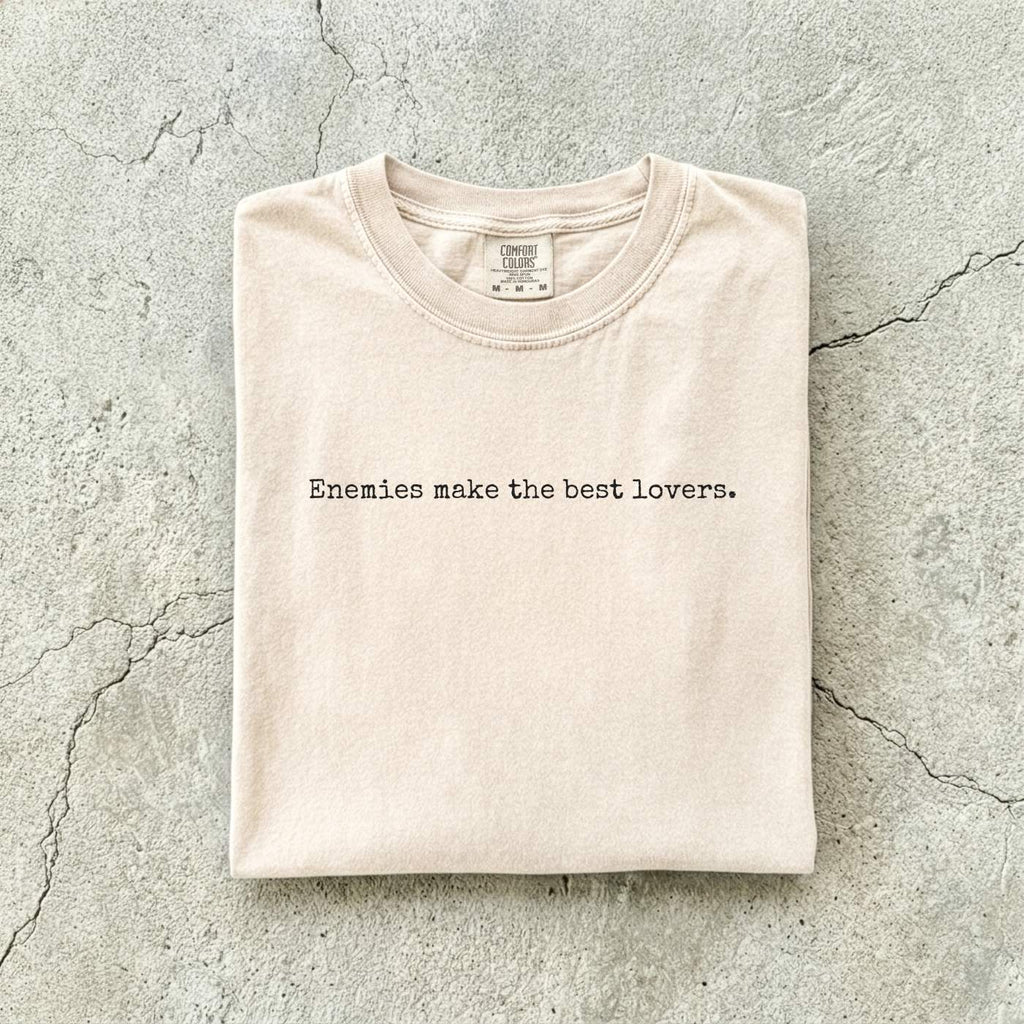 Ivory Comfort Colors tee folded flat on a concrete background reading "Enemies make the best lovers." in a minimal typewriter font.