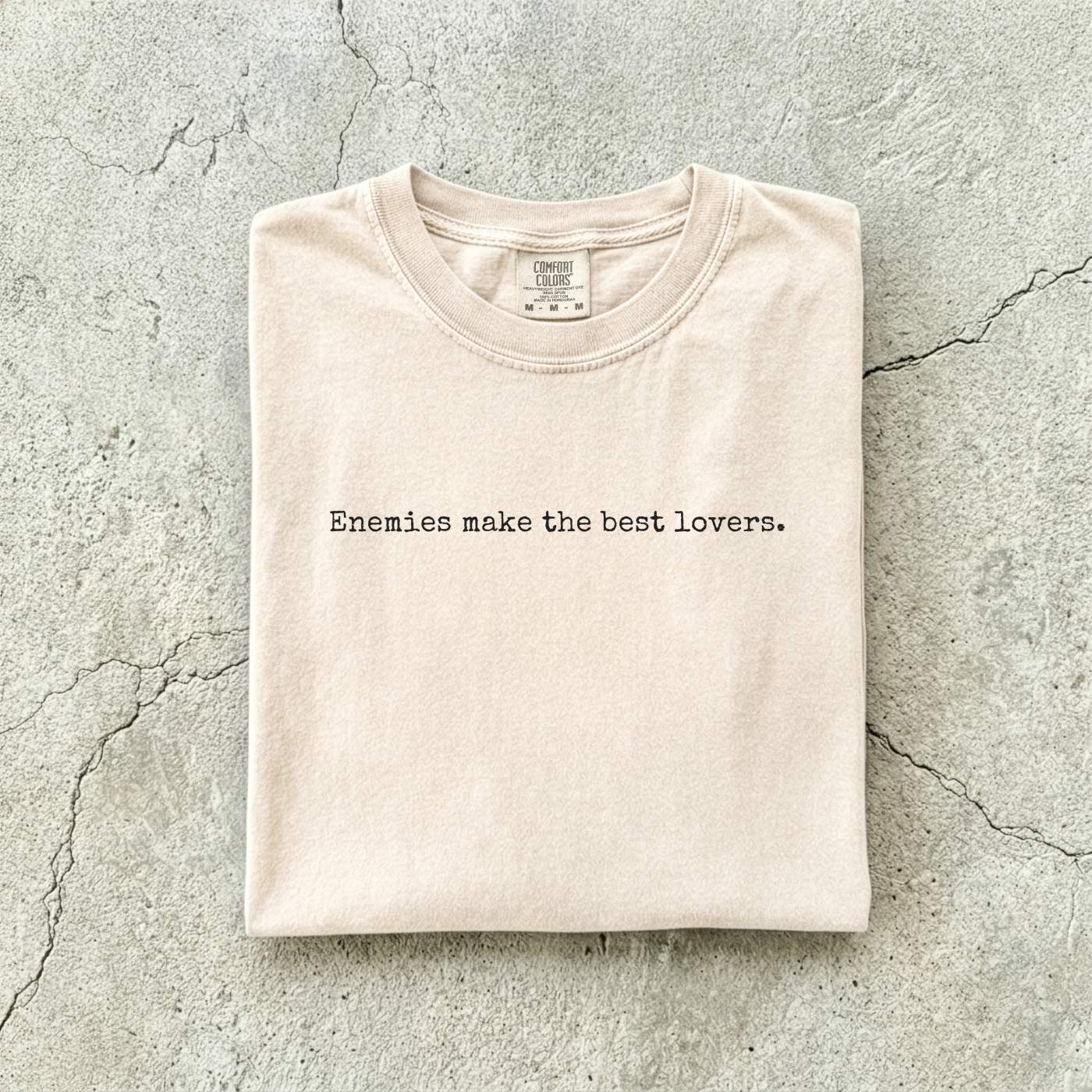 Ivory Comfort Colors tee folded flat on a concrete background reading "Enemies make the best lovers." in a minimal typewriter font.