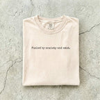 Ivory Comfort Colors tee folded flat on a concrete background reading "Fueled by anxiety and smut." in minimal typewriter font.