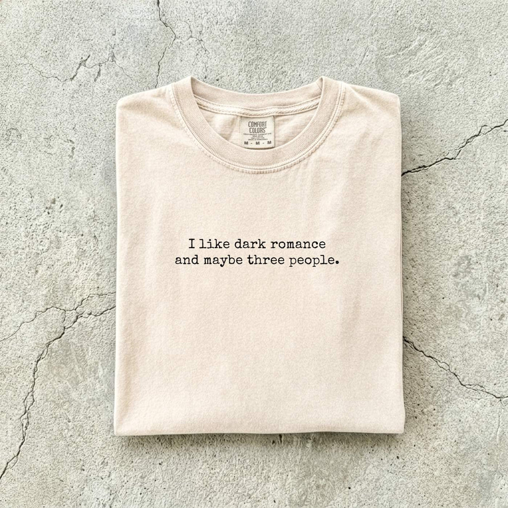 Ivory Comfort Colors tee folded flat reading "I like dark romance and maybe three people." in a minimalist typewriter font.