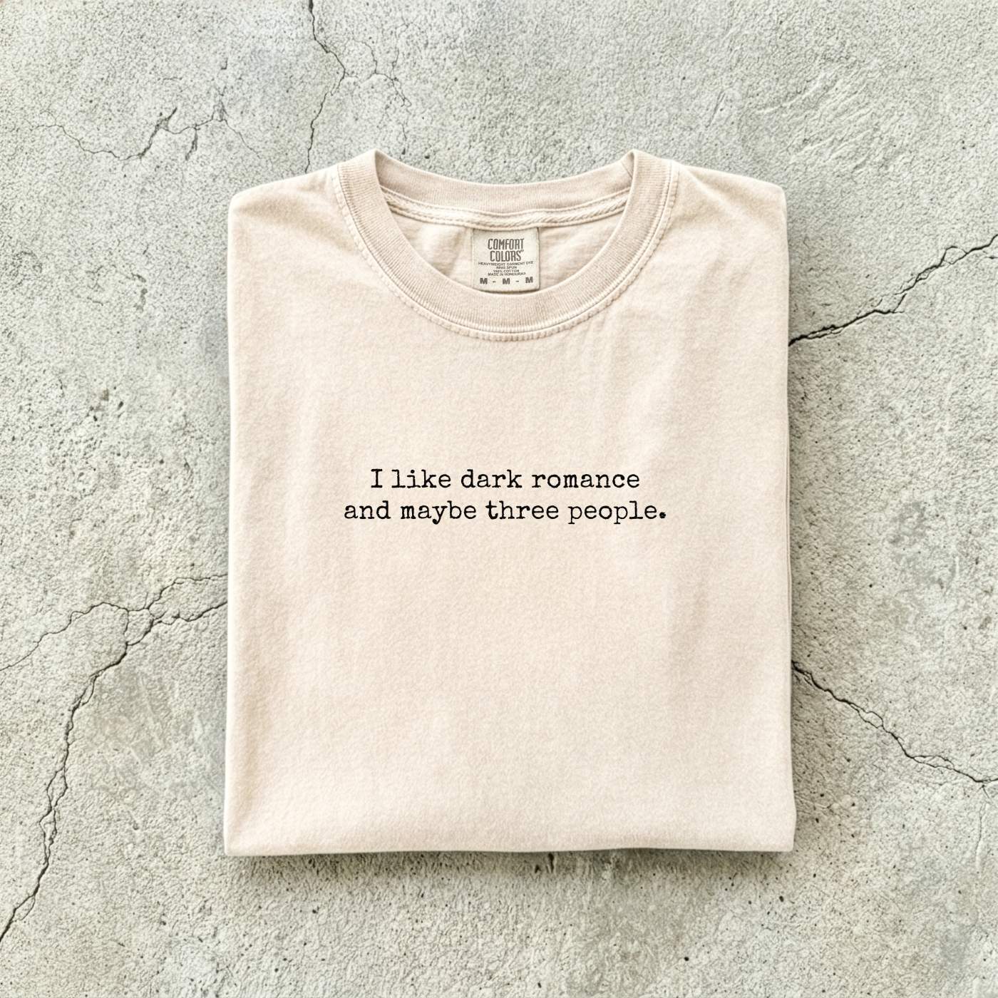 Ivory Comfort Colors tee folded flat reading "I like dark romance and maybe three people." in a minimalist typewriter font.